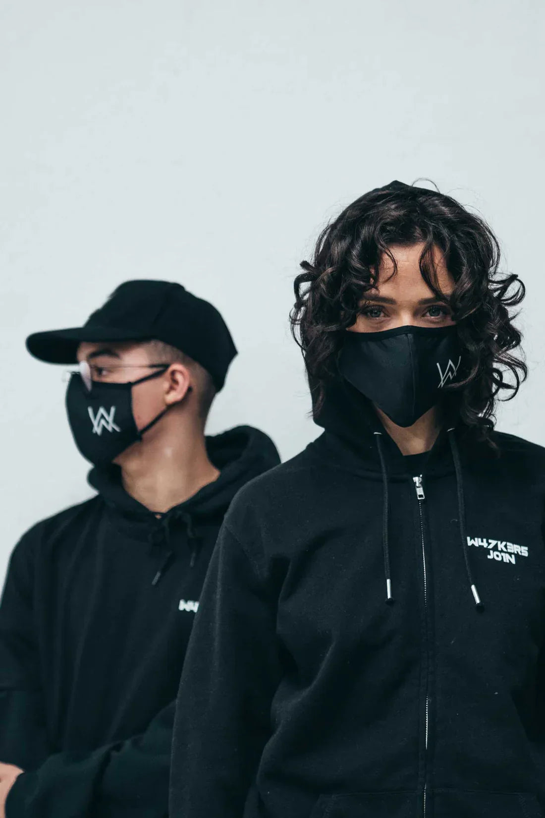 Two people showcasing Alan Walker merchandise, with one wearing a black 'AW' logo face mask and hoodie, and the other in a cap and hoodie, both embodying the urban style of the Walker Wear collection.