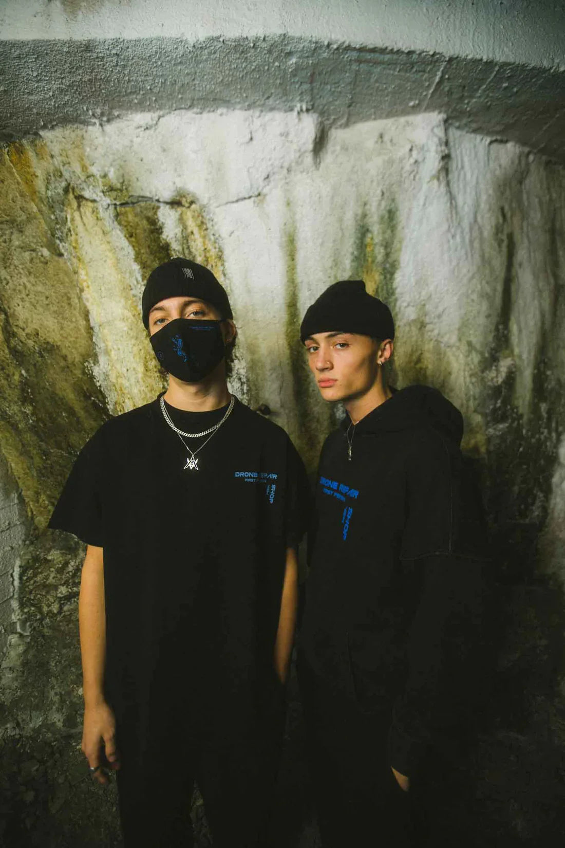 Two individuals standing side by side against a textured wall, one wearing a black Drone Repair Shop Mask and T-shirt, and the other in a hoodie, both with blue logo detailing, complemented by black beanies.