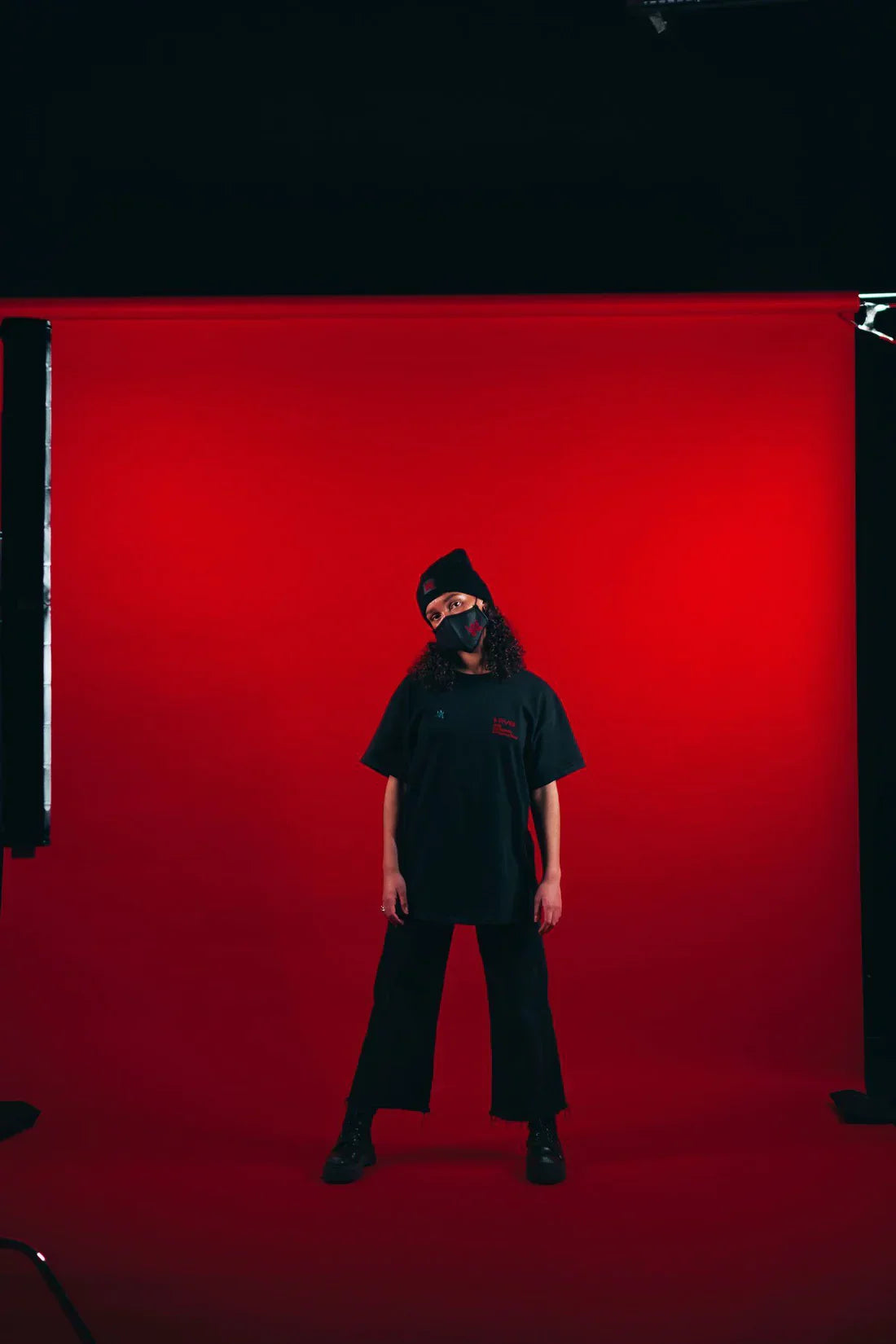 Individual standing in a photography studio setup with a striking red background, wearing a black AVI-8 t-shirt with red branding, black frayed jeans, black boots, and a beanie, complemented by a face mask with a red logo.