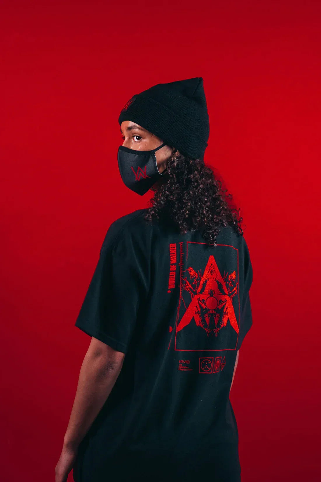 Individual from a side angle, wearing a black AVI-8 t-shirt with a large red graphic print on the back, a black beanie, and a face mask featuring the Alan Walker logo, set against a red background.