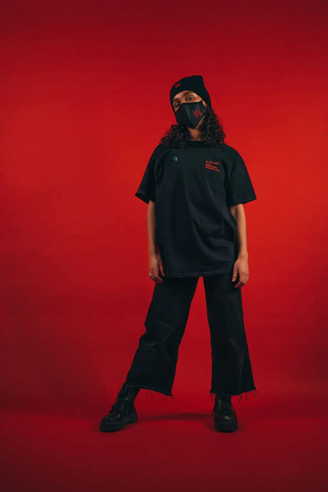 Individual posing against a red background, wearing a black AVI-8 t-shirt with subtle red branding on the chest, a black beanie with a logo, black frayed jeans, black boots, and a matching face mask.