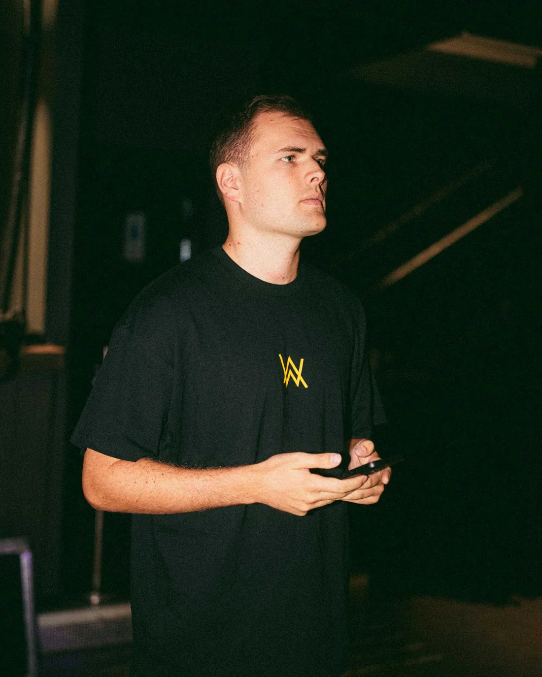 Man standing and looking to the side, holding a smartphone in his hands, wearing a black t-shirt with a small golden Alan Walker logo on the chest, in a dimly lit room.