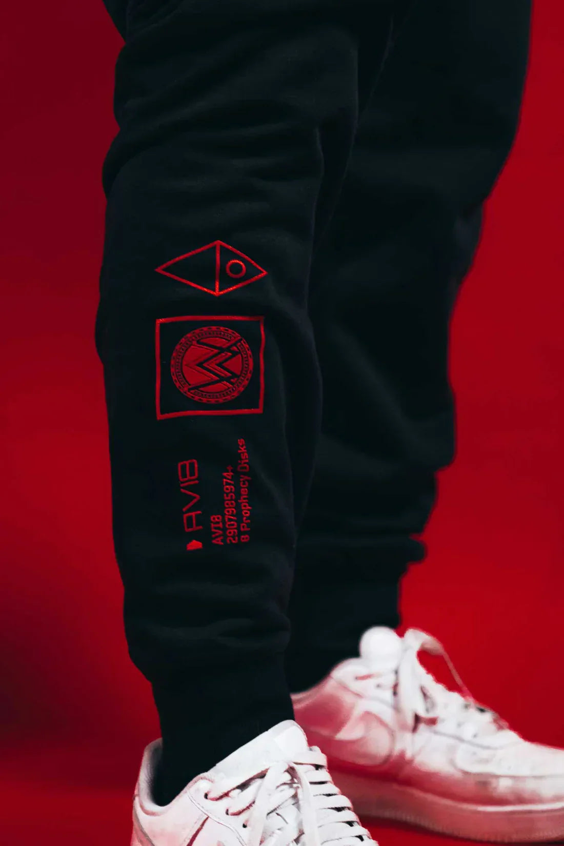 Close-up side view of a person's leg wearing black Alan Walker AVI-8 sweatpants with red graphics and text detailing down the side, paired with white sneakers, against a vivid red background.