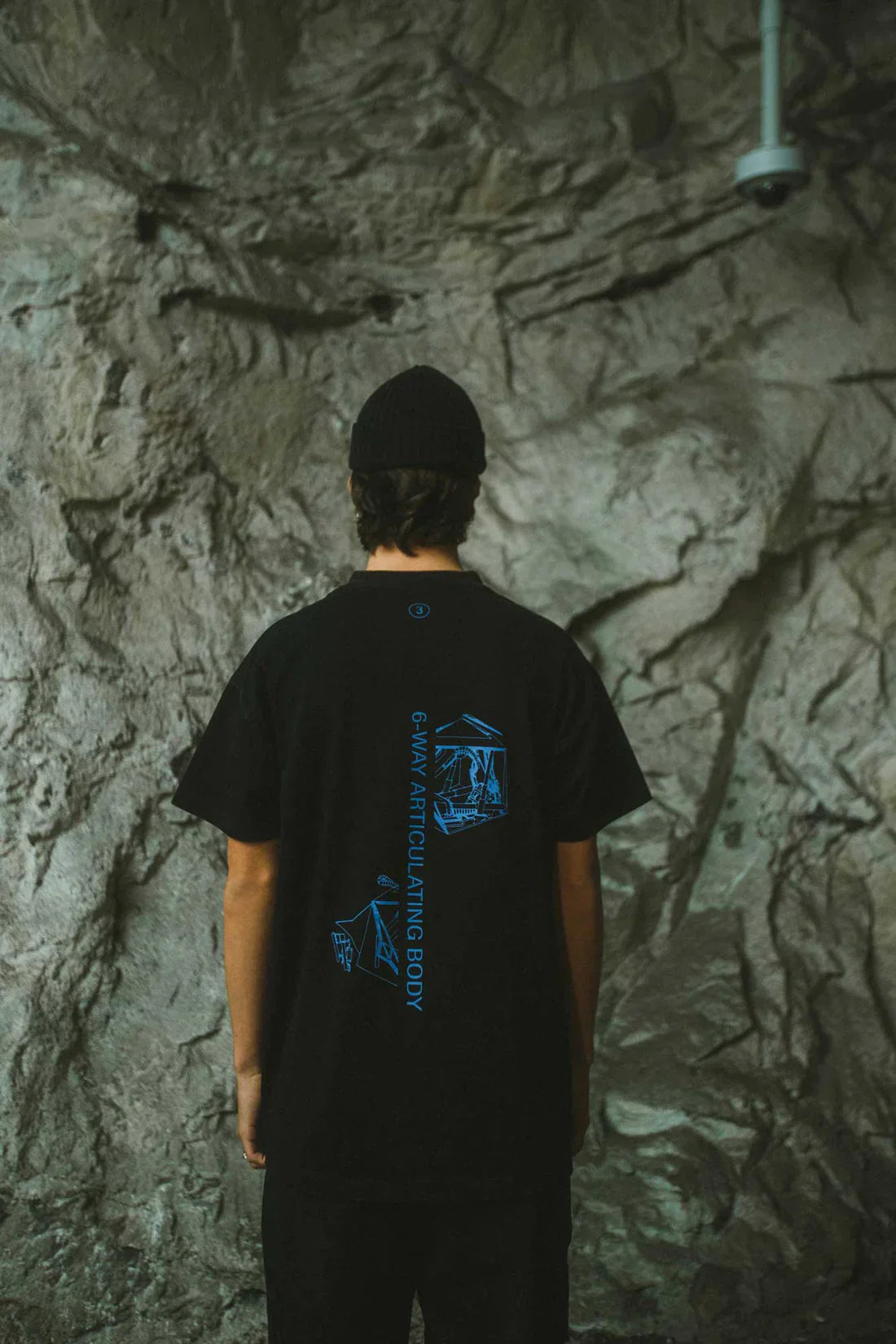Rear view of a person in a black beanie, wearing Alan Walker's Drone Repair Shop T-shirt with blue drone graphic on the back, set against a rugged concrete wall.