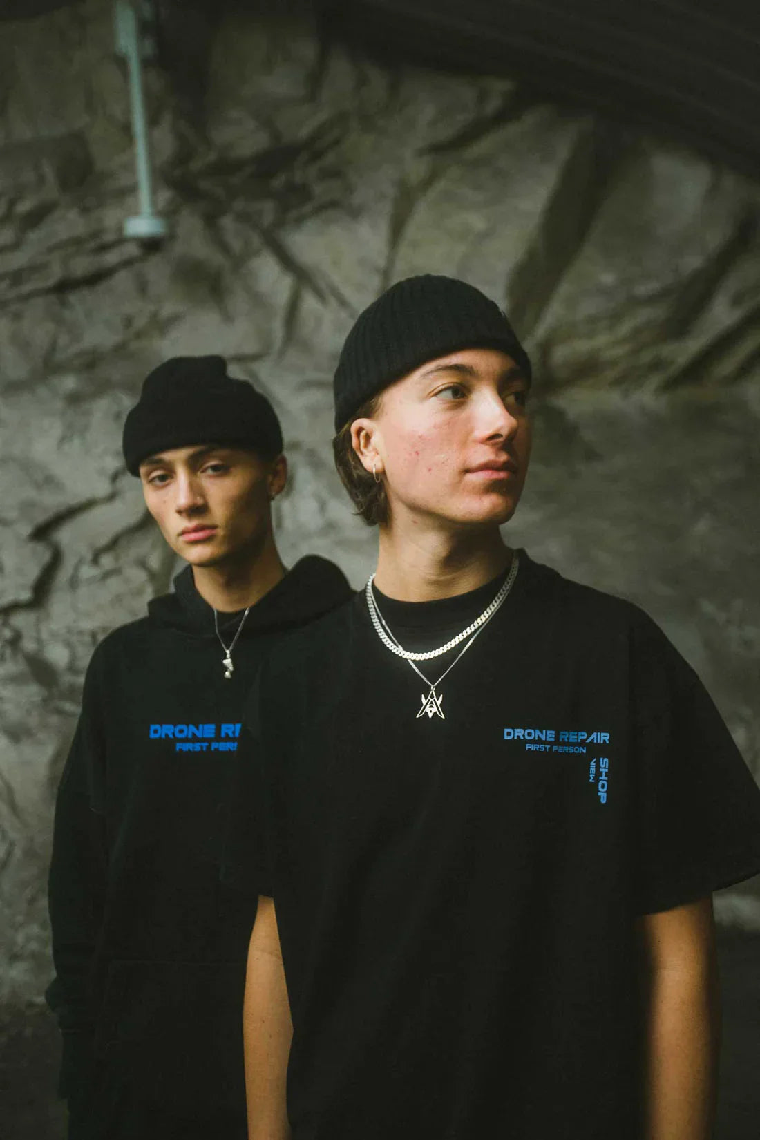 Two individuals wearing black beanies, with one in the Drone Repair Shop T-shirt and the other in a matching hoodie, both featuring blue logos, against an industrial concrete wall backdrop.