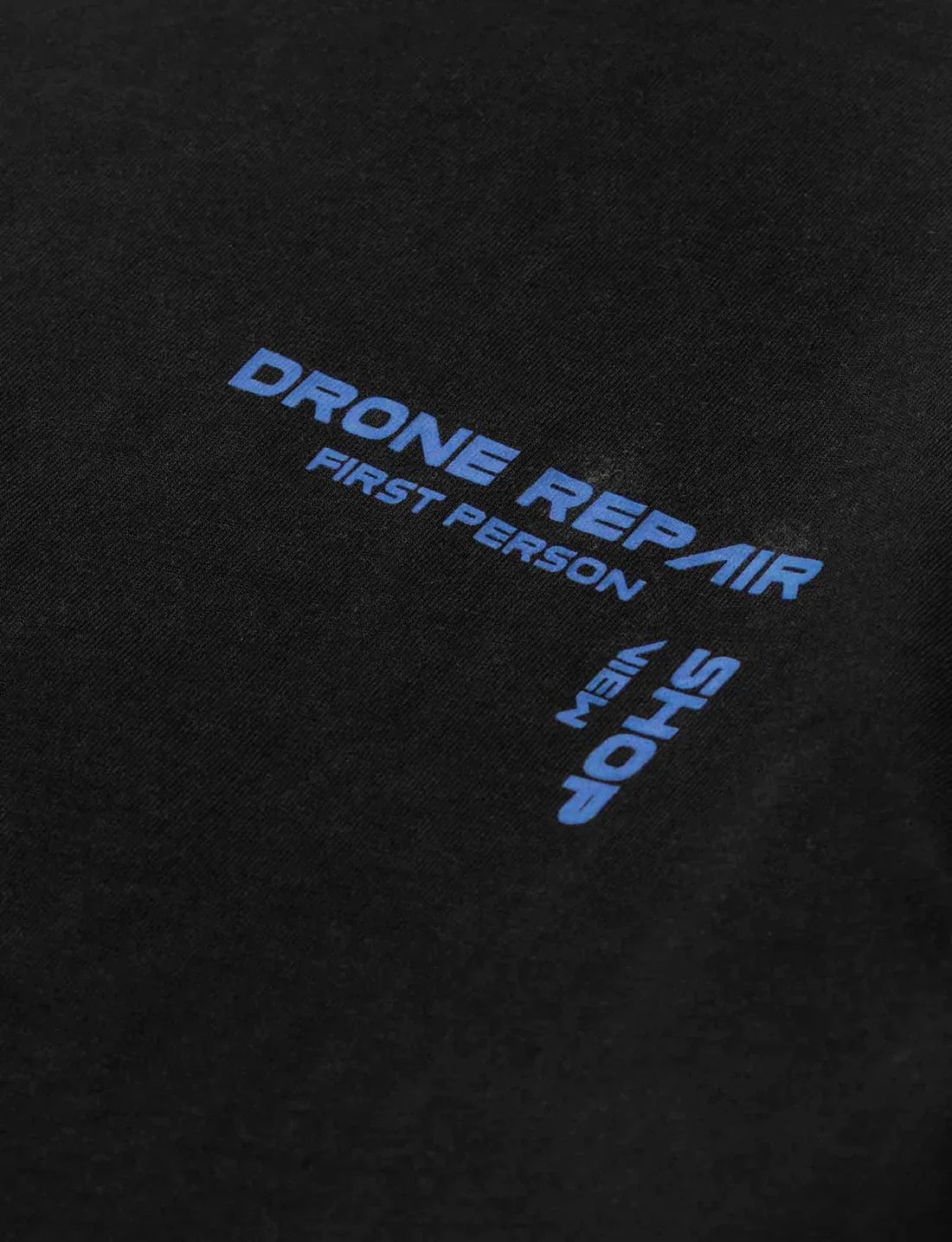 Detailed view of the front left chest area of Alan Walker's black Drone Repair Shop T-shirt, showing the blue logo with 'First Person Shop' text.