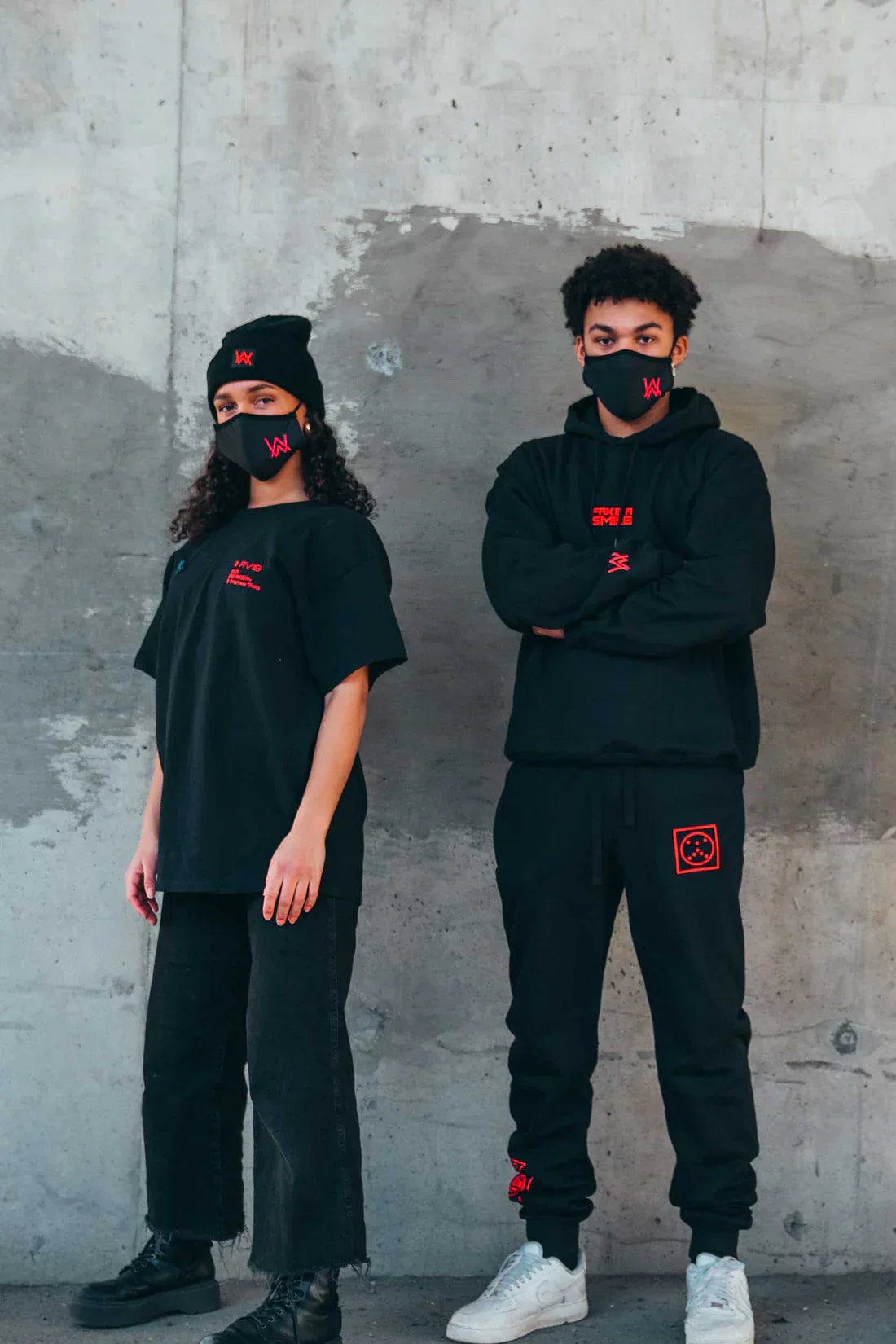 Two individuals posing against a concrete wall, one wearing a black hoodie and sweatpants with red Alan Walker branding, the other in a black t-shirt with similar branding, both wearing face masks with the Walker logo.