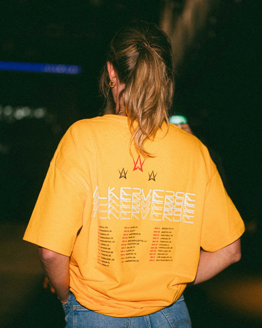 Person seen from behind wearing a yellow t-shirt with 'Walkerverse' in white lettering and a list of tour dates in orange, along with red Alan Walker logos, against a blurred urban night backdrop.