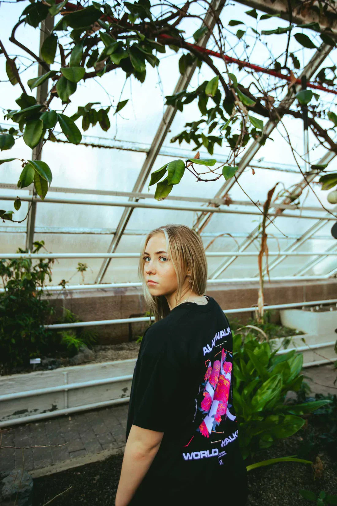 Female model elegantly sports the Melting Rose T-shirt in a lush greenhouse, blending style with nature.