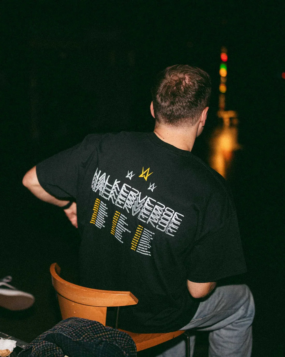 Man viewed from behind, wearing a black t-shirt with the 'Walkerverse' logo and tour dates in white and gold on the back, looking out into the night with city lights blurred in the distance.