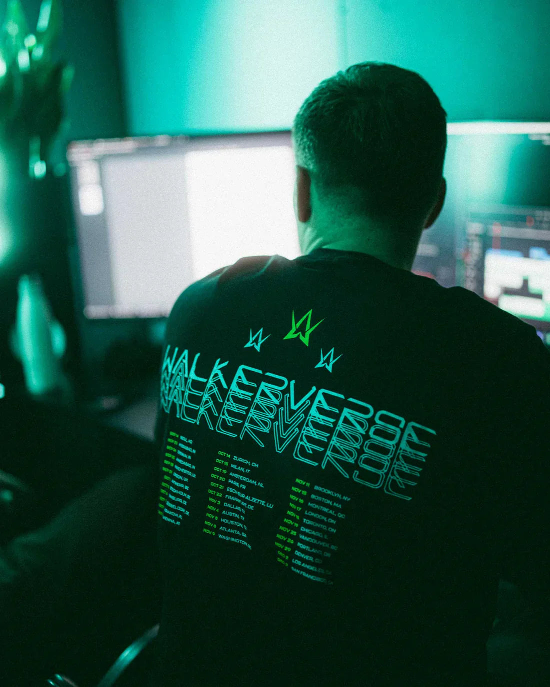Man seen from the back wearing a black t-shirt with 'Walkerverse' and tour dates printed in green and white, sitting in front of a computer screen with a blurred background.