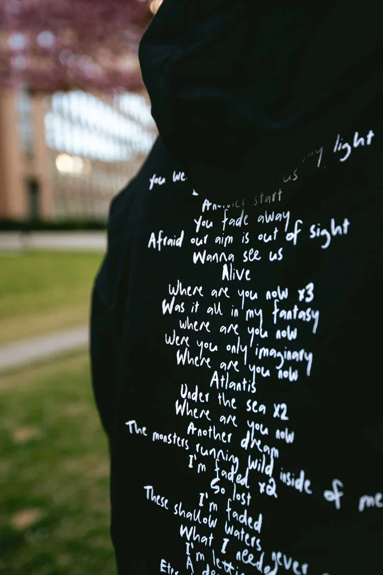 Close-up on the lyrics printed on the back of the Faded 2.0 Hoodie by Alan Walker, worn by a person with a blurred cityscape in the background.