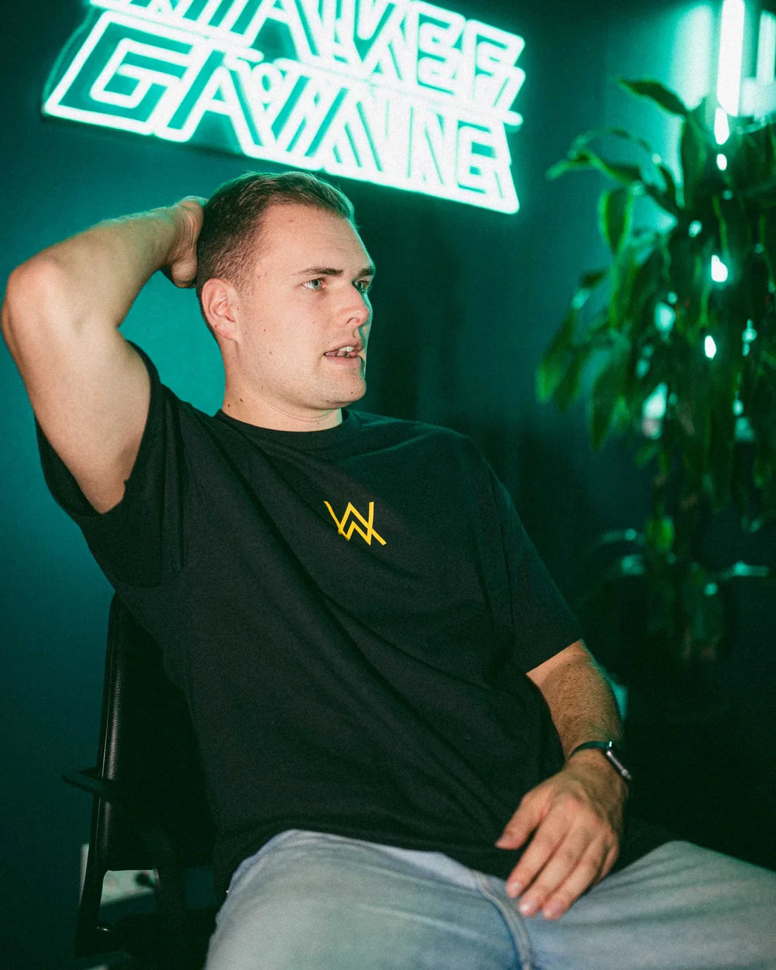 Man seated casually, wearing a black t-shirt with a golden Alan Walker logo, with a neon sign reading 'Walkerverse Gaming' in the background.