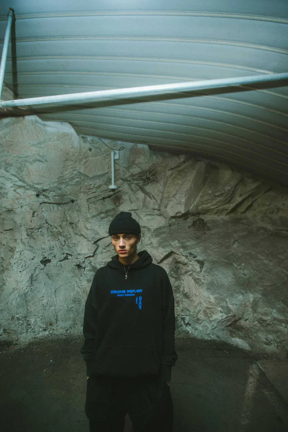 Person wearing a black Drone Repair Shop Hoodie with blue text, paired with black pants and a beanie, standing against a textured concrete background in an industrial setting.