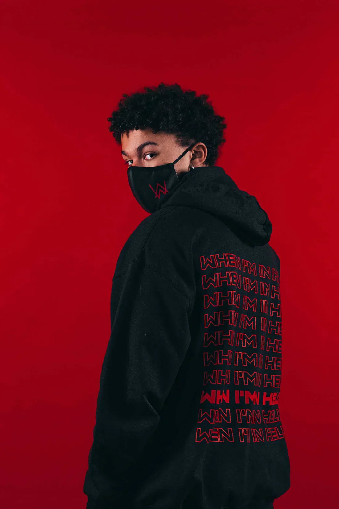 Side view of a person modeling the Alan Walker Fake a Smile Hoodie with the red 'WHEN I'M IN HELL' text visible along the arm, set against a bold red background.