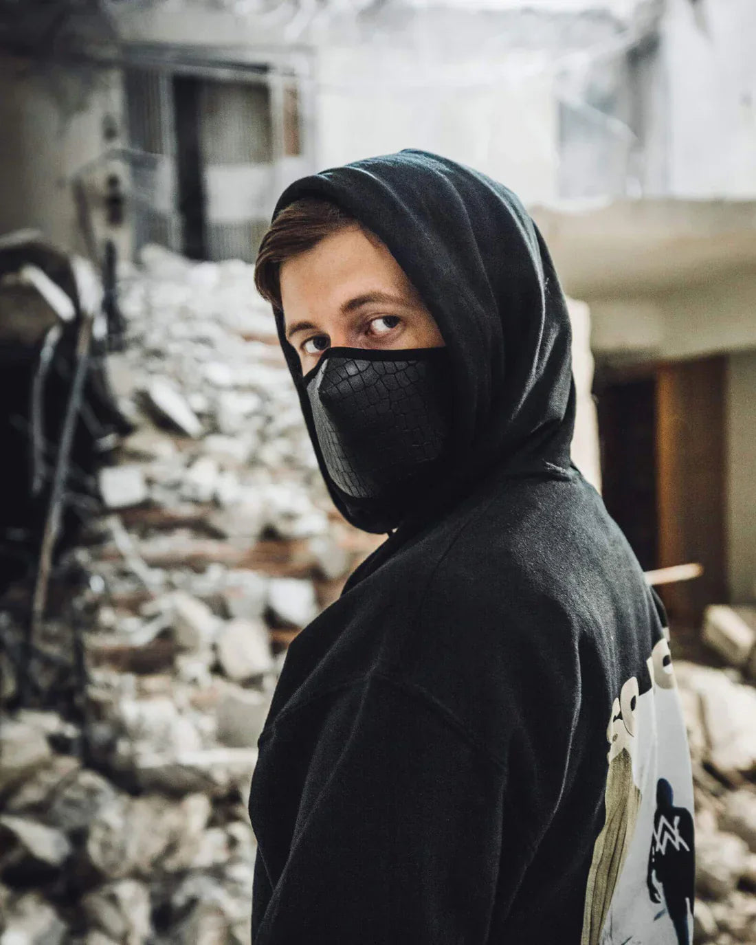 Individual wearing Alan Walker's Faded black hoodie with hood up and a black face mask, turning towards the camera with a backdrop of urban decay, highlighting the theme of the 'Faded' series.